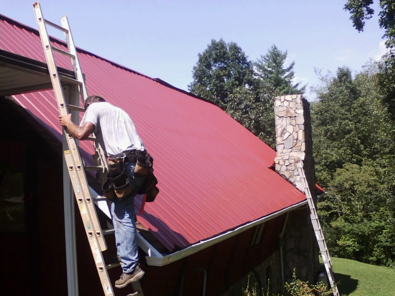 Roofer on a ladder beside a red metal roof and stone chimney — Soffit & Fascia Repair by Roofing Brothers in Walpole